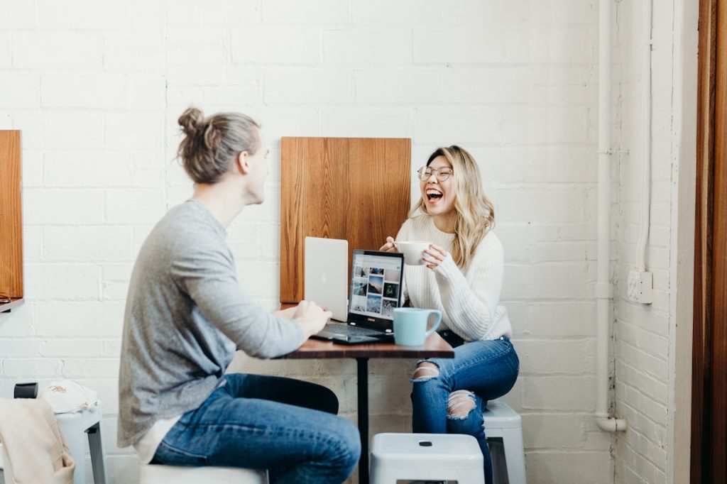 A man and a woman laughing whilst sitting at a desk with their laptops.